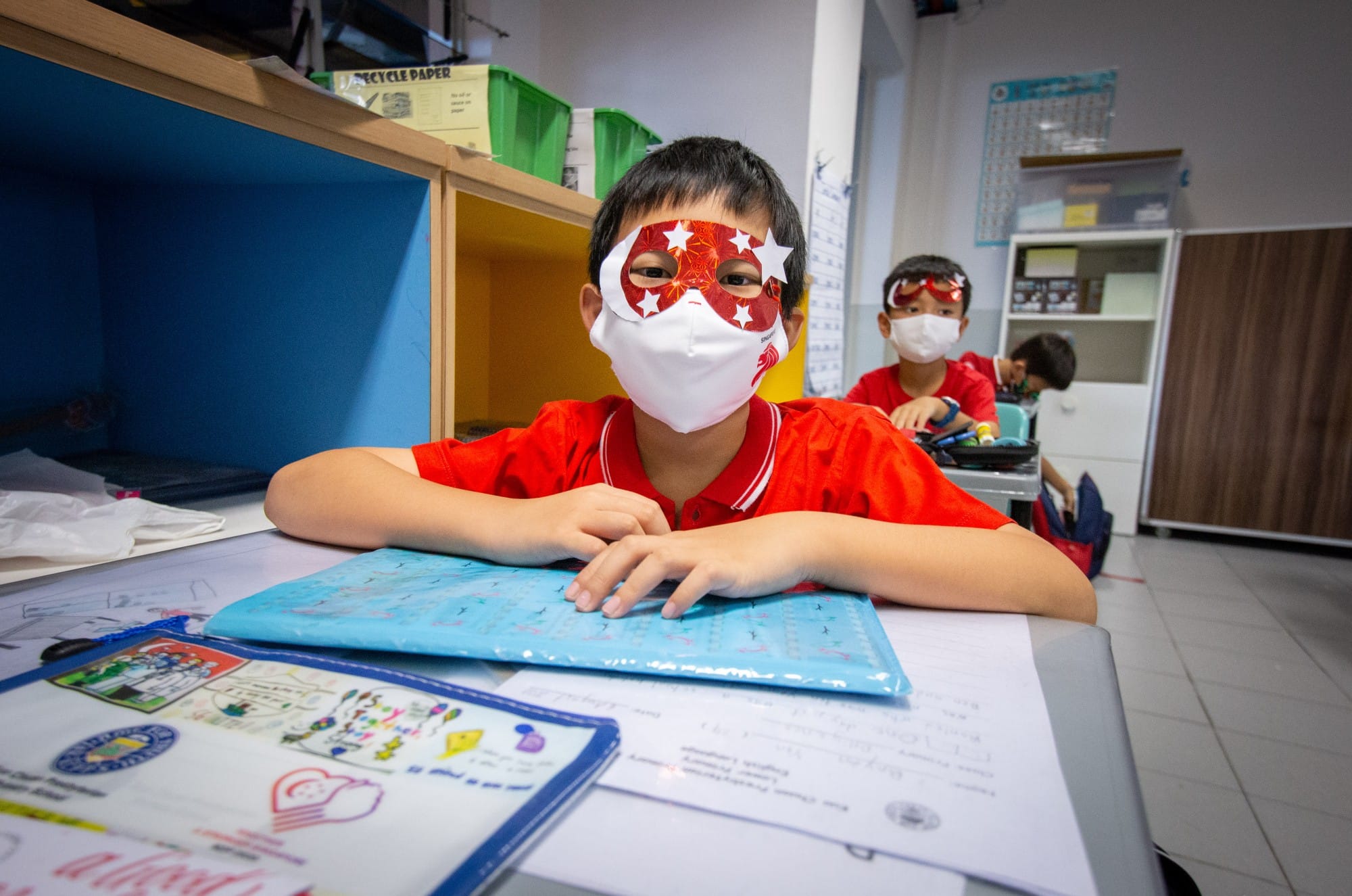 A young boy in a red shirt wearing a face mask and red star-patterned paper mask sits at a classroom desk.
